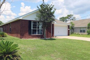 A single-story brick house with a white garage door, a small front yard with grass and a tree, and a clear sky. - Celebrate Mother`s Day on the Beach (Navarre)