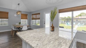 A modern kitchen with a large marble island, a dining area with a table and chairs, and large windows with blinds. - Celebrate Mother`s Day on the Beach (Navarre)