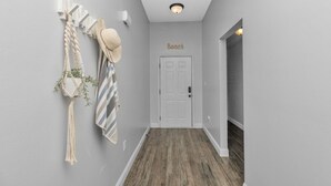 A hallway with a wooden floor, a white door with a 'Beach' sign above it, and a macrame wall hanging with a hat and greenery. - Celebrate Mother`s Day on the Beach (Navarre)