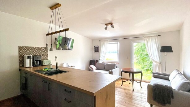 Eine moderne Küche mit einer zentralen Kochinsel, einem Essbereich mit Sofa und Tisch sowie Blick auf Grünflächen durch ein Fenster.
