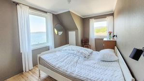 A bedroom with a bed, a chair, a desk, and a view of the sea.