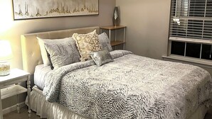 Ein Schlafzimmer mit einem großen Bett, einem Nachttisch, einer Lampe, einem Fenster mit Jalousien und einem Wandbild.