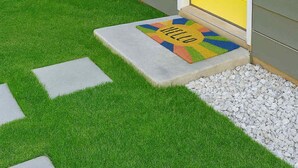 Ein Haus mit gelber Haustür, einer kleinen Veranda und einer Fußmatte mit dem Aufdruck 'HELLO', sowie ein Holzzaun.