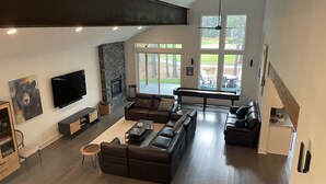 Un salon spacieux avec un grand canapé d’angle, un téléviseur à écran plat fixé au mur, une cheminée et une vue sur l’extérieur grâce à de grandes fenêtres.