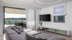 Een moderne woonkamer met een grijze hoekbank, een flatscreen televisie aan de muur en uitzicht op een balkon met tuinmeubilair.