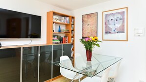 Ein modernes Wohnzimmer mit einem Glastisch, weißen Stühlen, einer Bücherwand und Kunstwerken an den Wänden.