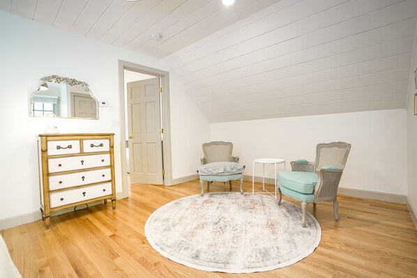 A room with a wooden floor, a round rug, a dresser with drawers, two chairs, a small table, and a mirror on the wall.