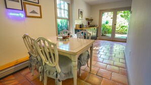 A dining area with a table and chairs, a kitchen in the background, and a view of greenery through the window. - Private Cape Cod Cottage. Walk to Ellis Landing Beach! (Brewster)
