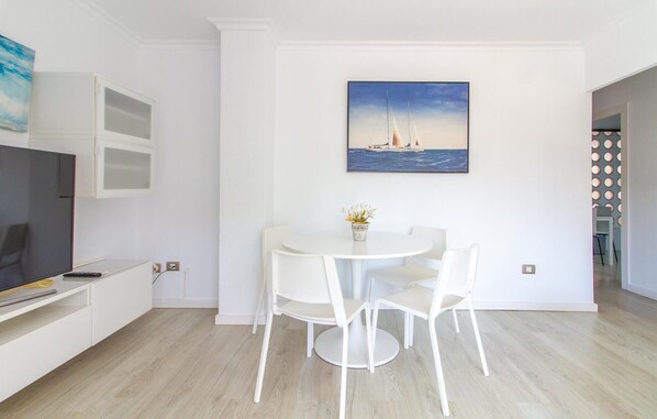 Un espace repas moderne et minimaliste, avec une table ronde et des chaises blanches, un tableau représentant un voilier accroché au mur et un téléviseur posé sur un meuble.