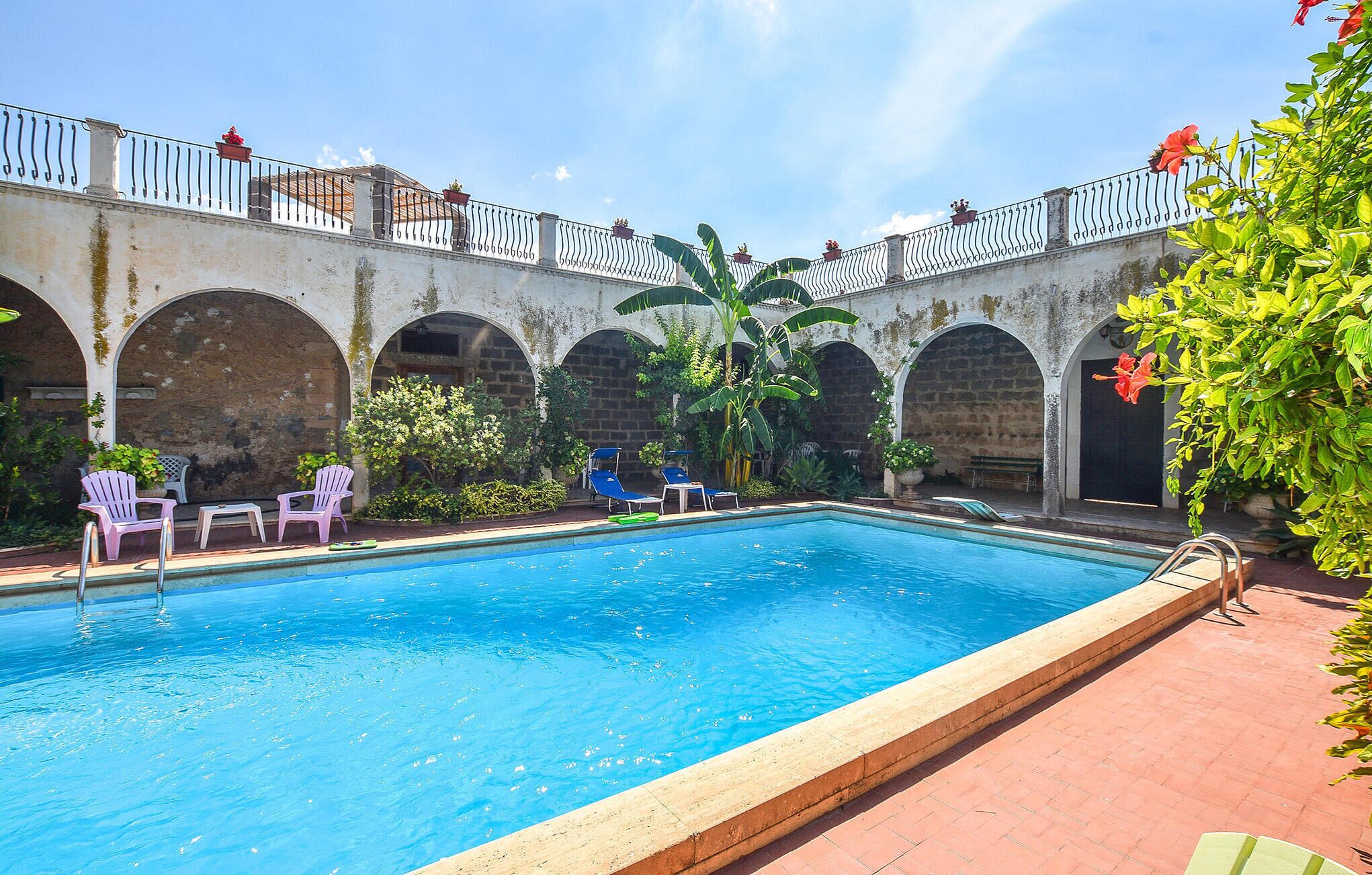 Una piscina con plataforma de baldosas, rodeada de muros de piedra en arco y una barandilla de metal.