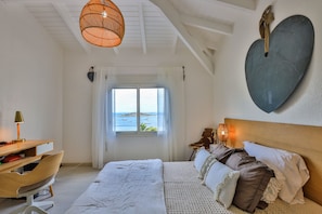 Quarto com cama, escrivaninha, cadeira e vista para o mar pela janela.