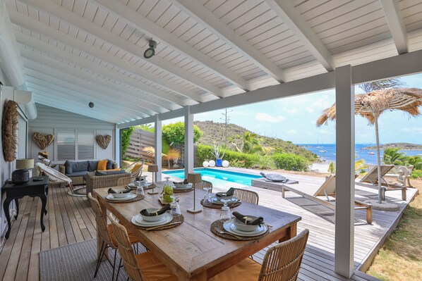 Uma área de refeições ao ar livre coberta, com uma mesa de madeira posta para quatro pessoas, com vista para uma piscina e uma paisagem marítima deslumbrante.