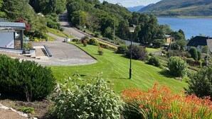 A residential area with a well-maintained lawn, a small building with a porch, and a view of a lake and hills in the background. - FOINAVEN HOUSE, Quality 4 star self catering accommodation with panoramic views. (Ullapool)
