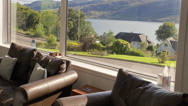 A room with a large window offering a view of a lake and mountains. There is a leather sofa with cushions and a small table with a book and a cup. - FOINAVEN HOUSE, Quality 4 star self catering accommodation with panoramic views. (Ullapool)