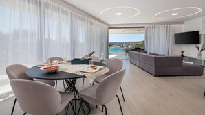 Ett modernt matområde med ett bord dukat för två personer, en soffa och en TV i ett vardagsrum med utsikt över havet.