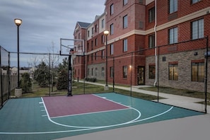 Una cancha de baloncesto al aire libre junto a un edificio de ladrillo con varias ventanas y una valla.