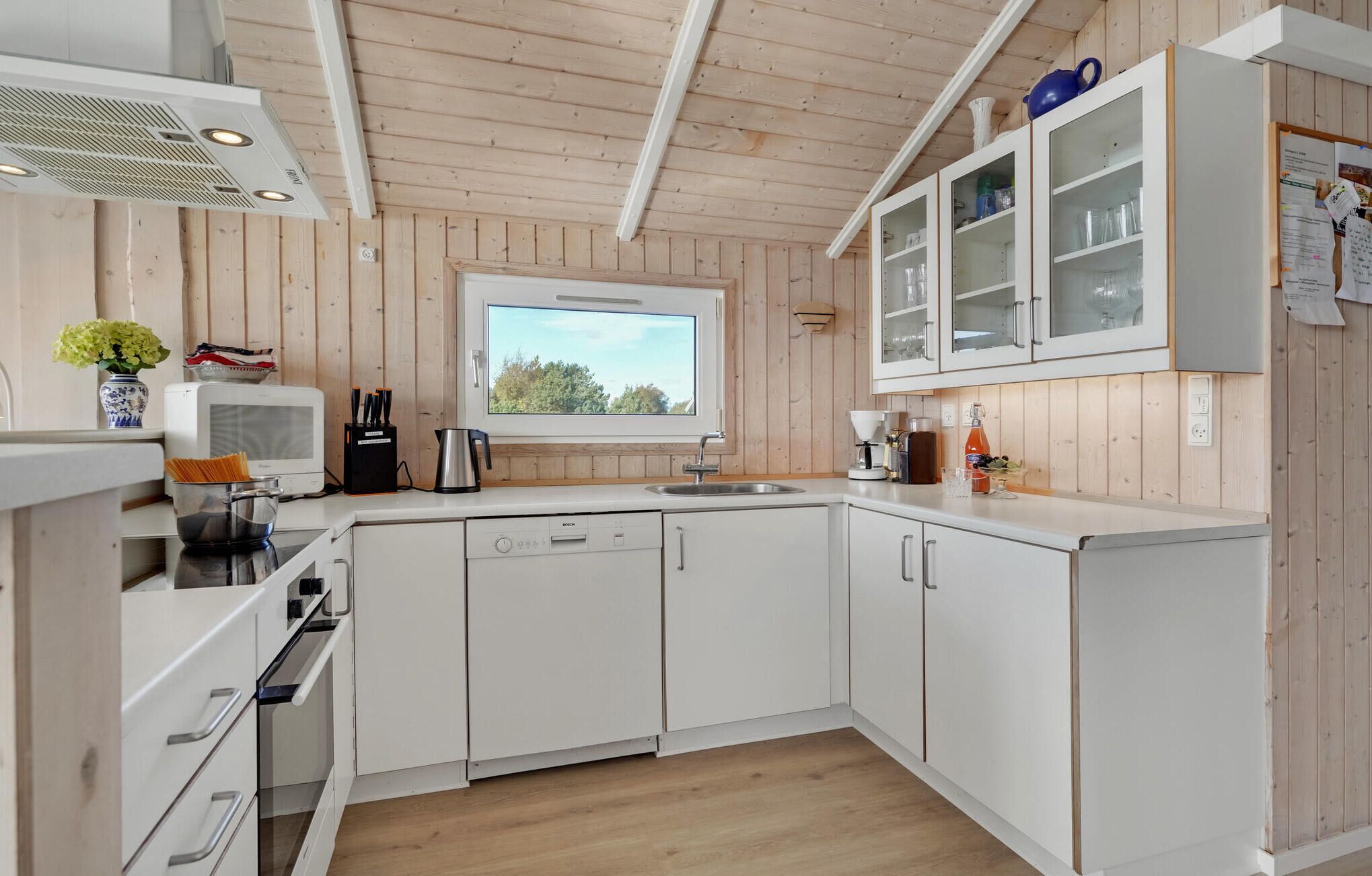 A modern kitchen with white cabinets, a Bosch dishwasher, a window with a view of trees, and wooden walls.