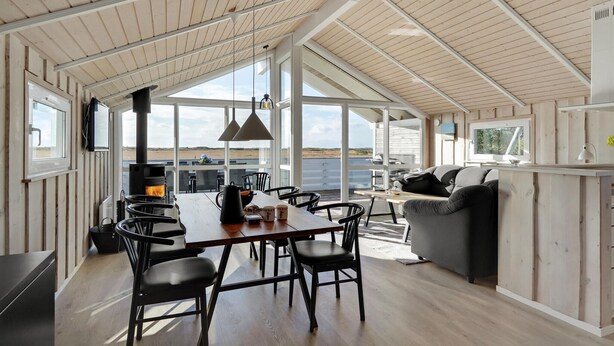 A spacious living area with a large wooden table, black chairs, and a grey sofa. A fireplace is visible in the background, and the room has large windows offering a view of the beach. - Gorgeous home in Rømø with sauna (Rømø)