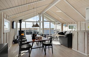 A spacious living area with a large wooden table, black chairs, and a grey sofa. A fireplace is visible in the background, and the room has large windows offering a view of the beach. - Gorgeous home in Rømø with sauna (Rømø)