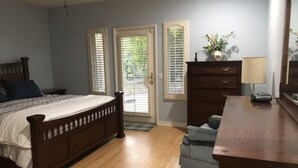 A bedroom with a wooden bed, a dresser, and a chair. There are two windows with blinds, a ceiling fan, and a door leading outside.