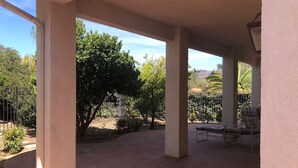 A covered patio area with brick flooring, a metal chair, and a view of a garden with trees and a fence.