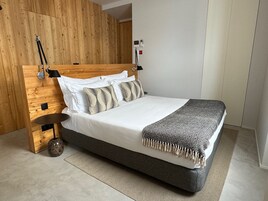 A modern bedroom with a wooden headboard, white bedding, patterned pillows, and a grey blanket.