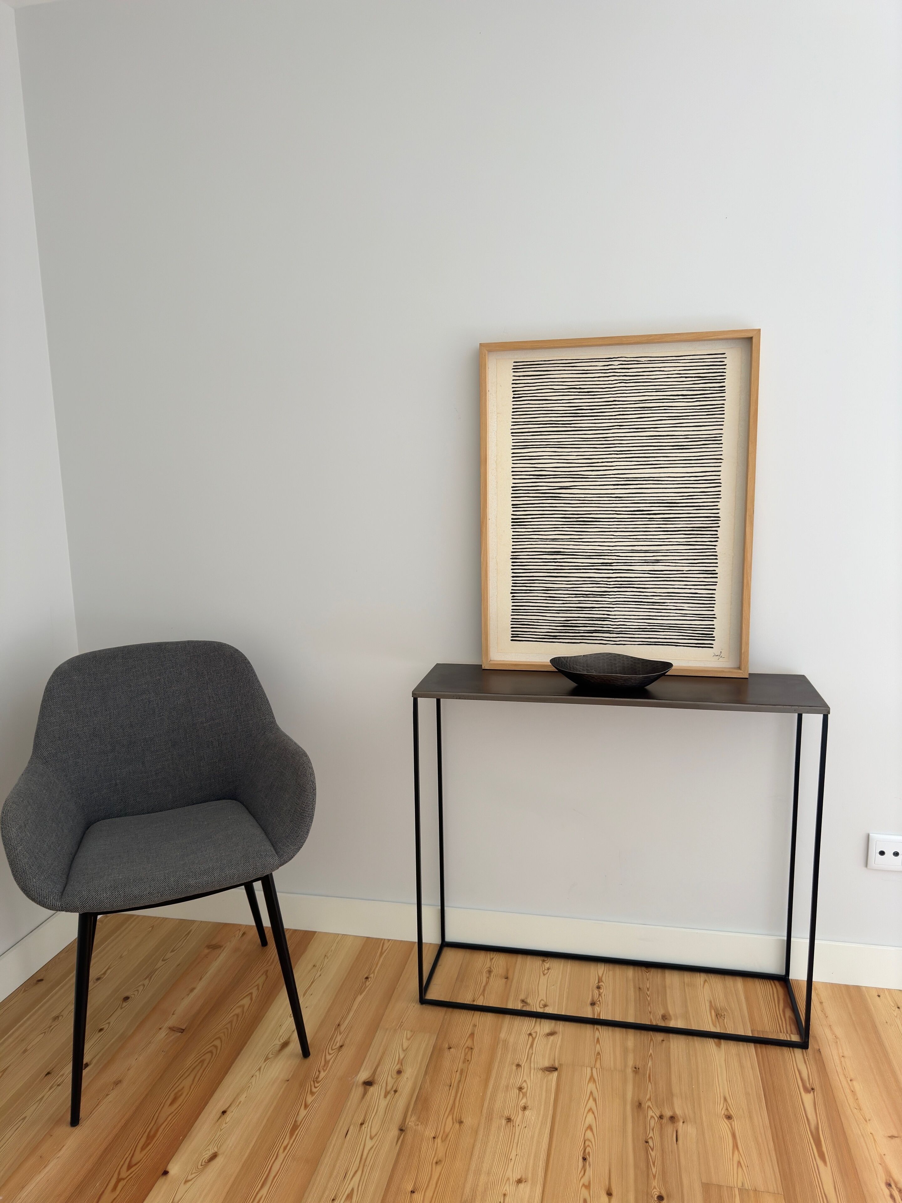 A modern room with a grey armchair, a wooden floor, a black-framed table, and a framed abstract artwork.