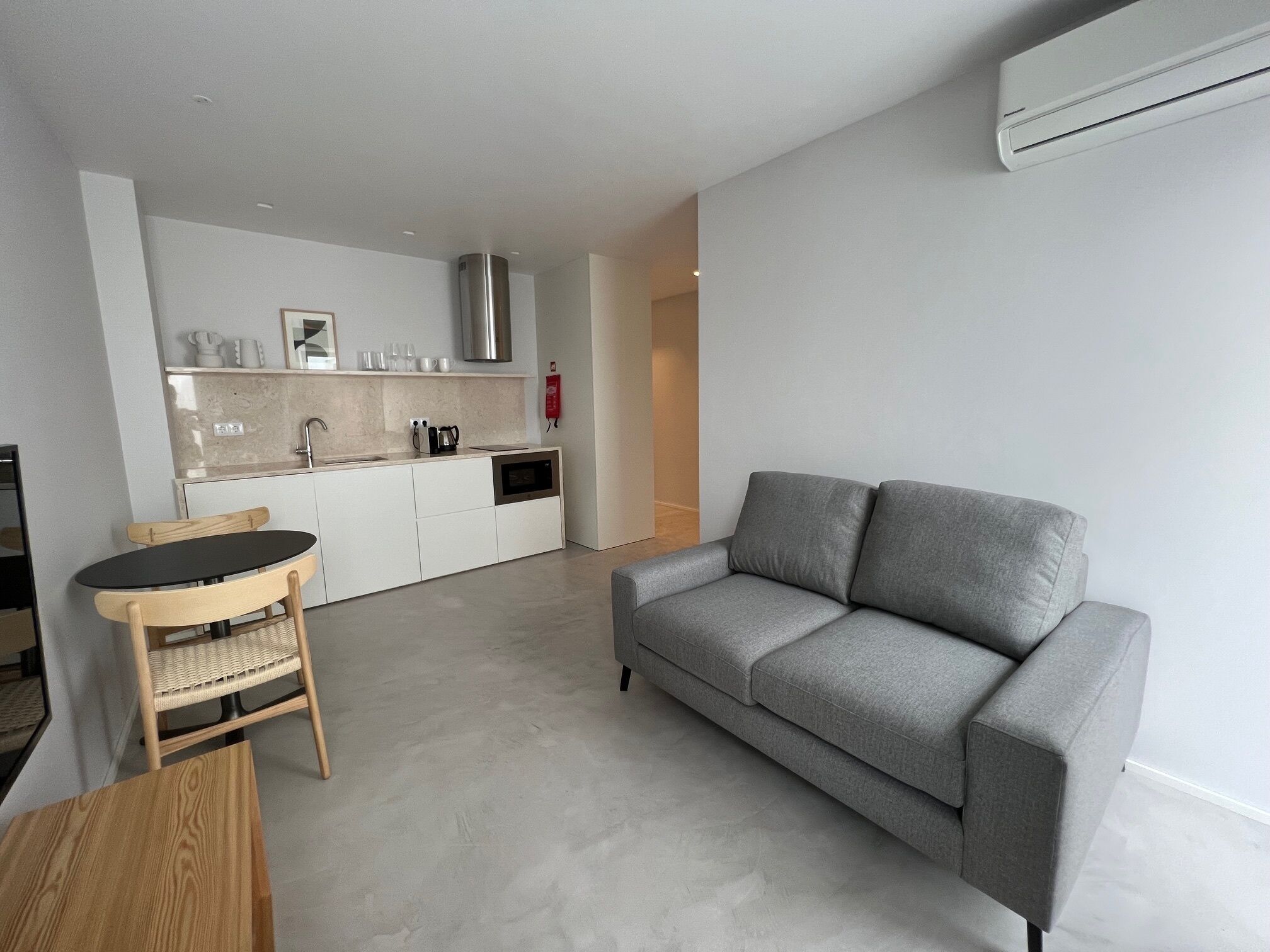 A modern living room with a grey sofa and a small dining table with chairs. A kitchen with white cabinets and a stainless steel range hood is visible in the background.