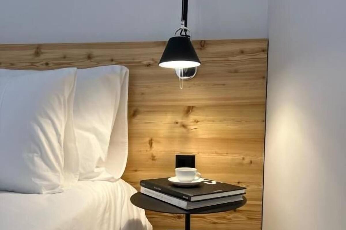 A bedroom with a wooden headboard, a bed with white bedding, a small round table with a lamp, a cup, and books.