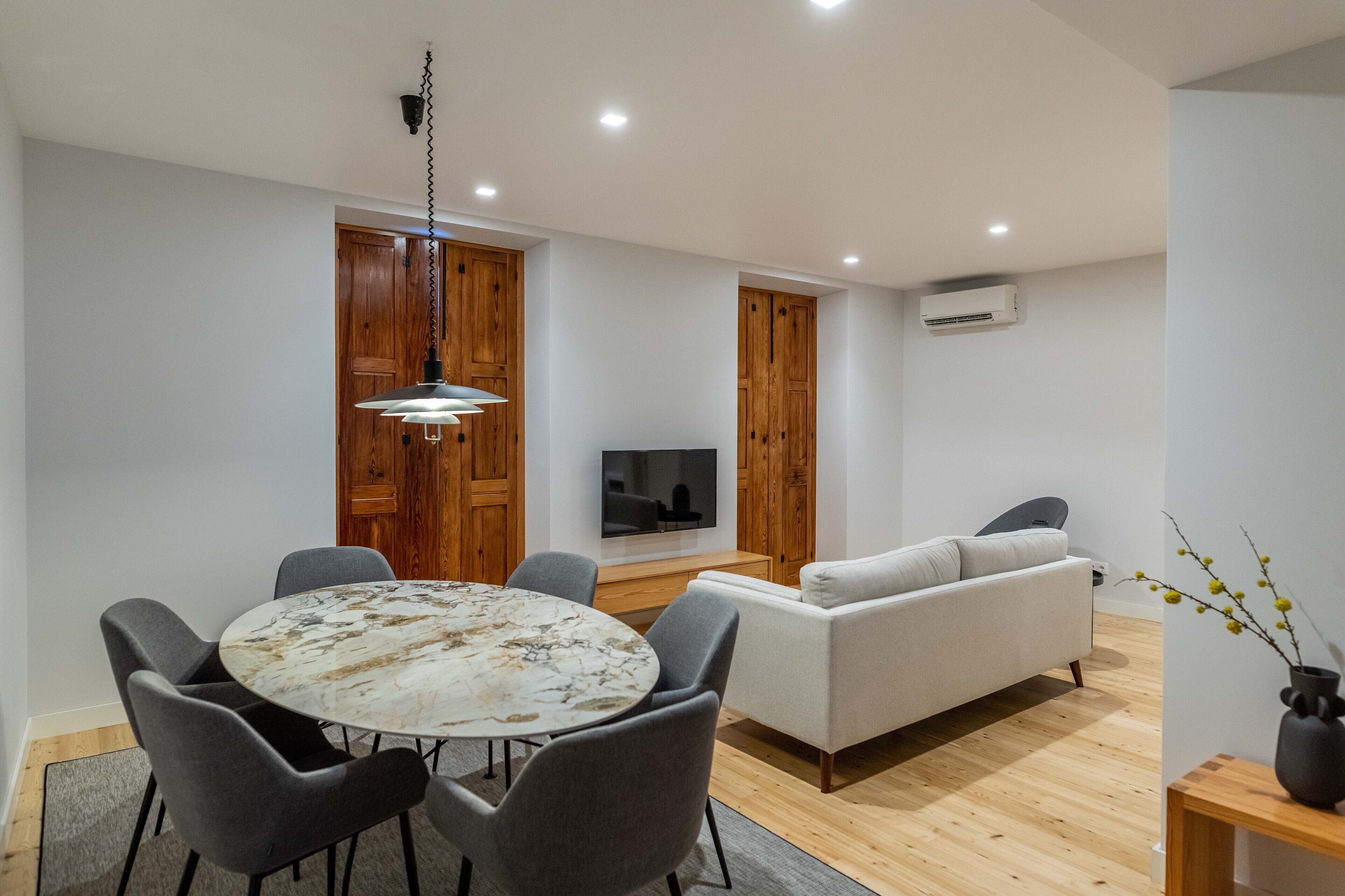 A modern living room with a round marble table, grey chairs, a white sofa, and a TV mounted on the wall.