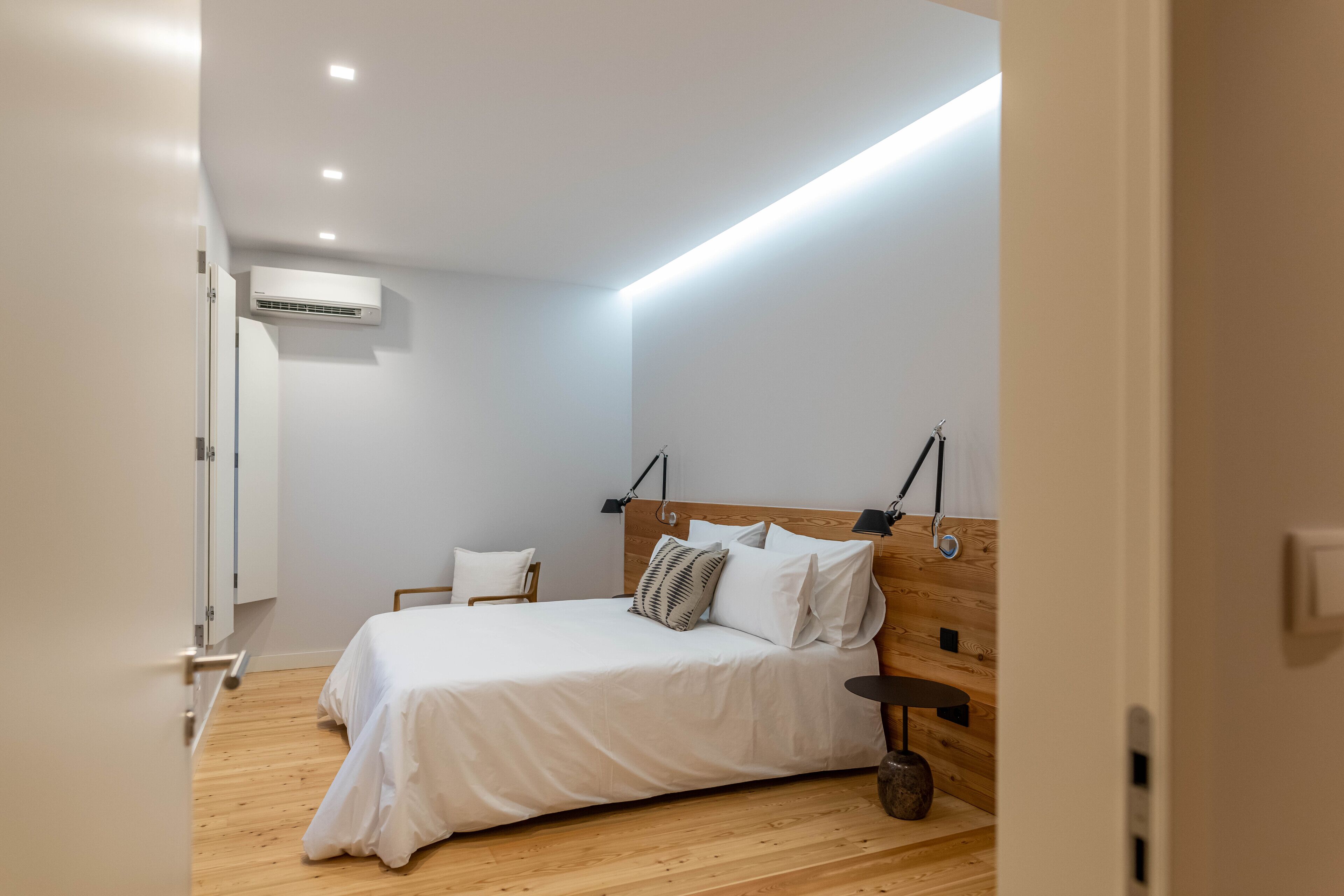Quarto moderno com piso de madeira, cama com roupa de cama branca, mesa de cabeceira e ar condicionado fixado na parede.