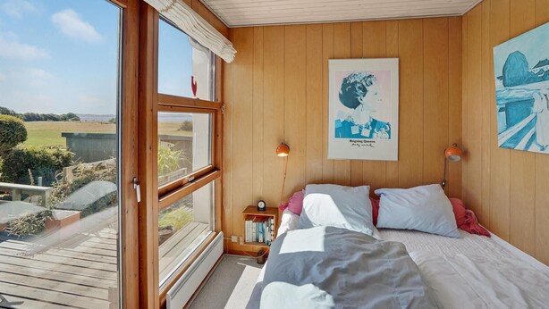 A bedroom with a large window, wooden walls, a bed with pillows, a bedside table with a lamp, and a poster on the wall. - Awesome home in Broager with WiFi (Broager)
