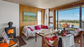 A cozy living room with a fireplace, a sofa with colorful cushions, a dining table, and a view of the countryside through large windows. - Awesome home in Broager with WiFi (Broager)