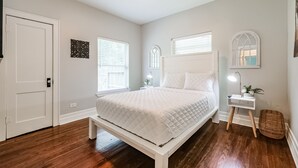 Une chambre avec un lit blanc, un plancher en bois, une lampe de chevet et une fenêtre avec des stores.