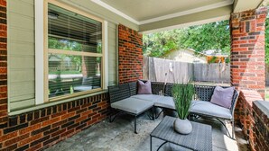 Un patio couvert avec un mur en briques, une fenêtre avec des stores et du mobilier d’extérieur comprenant un banc et une table.