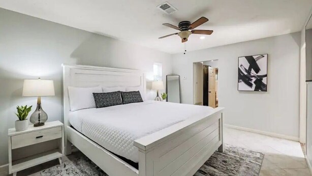 Une chambre à coucher moderne avec un lit blanc, un ventilateur de plafond, une table de chevet avec une lampe et une petite plante.