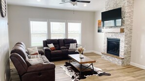 A modern living room with a stone fireplace, leather sofas, and a ceiling fan. - 3 Peaks Mountain View Getaway w/Hot Tub! (Cedar City)