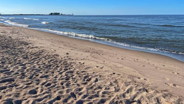 Una playa arenosa con huellas que se adentran en el agua, un cielo despejado y una estructura distante en el horizonte.