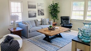 Una sala de estar con un sofá gris, una mesa de centro de madera, un sillón de cuero negro, una planta en maceta y un ventanal grande.