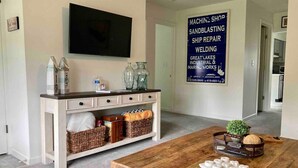 Una sala de estar moderna con una mesa de centro de madera, una consola blanca con cestas, un televisor de pantalla plana montado en la pared y un letrero publicitando servicios de granallado, reparación de embarcaciones y soldadura.