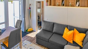 Ein modernes Wohnzimmer mit einem grauen Sofa, orangefarbenen Kissen, einem Esstisch mit Stühlen und einem Bücherregal mit Büchern und Dekogläsern.