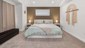 A bedroom with a bed, bedside tables, a window with blinds, a wall-mounted macrame piece, and a dark bench. - Luxury 3-bedroom Tri-Level w/ Game Room (Hoquiam)