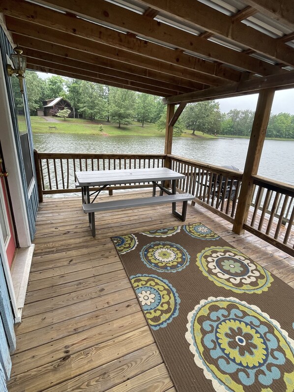 A wooden deck with a picnic table and benches, overlooking a lake with a grassy area and trees in the background.