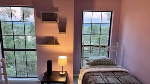 A bedroom with a bed, a nightstand, a lamp, and two windows offering a view of trees.