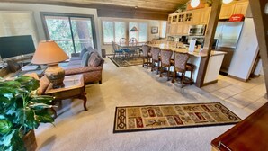 A spacious living room with a sofa, coffee table, and a TV. There is a dining area with chairs and a rug. The kitchen has wooden cabinets and a refrigerator. - Mountain Back (Mammoth Lakes)