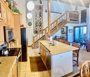 Une cuisine avec des meubles en bois, un four encastré et un four à micro-ondes. Un escalier en bois mène à une mezzanine.