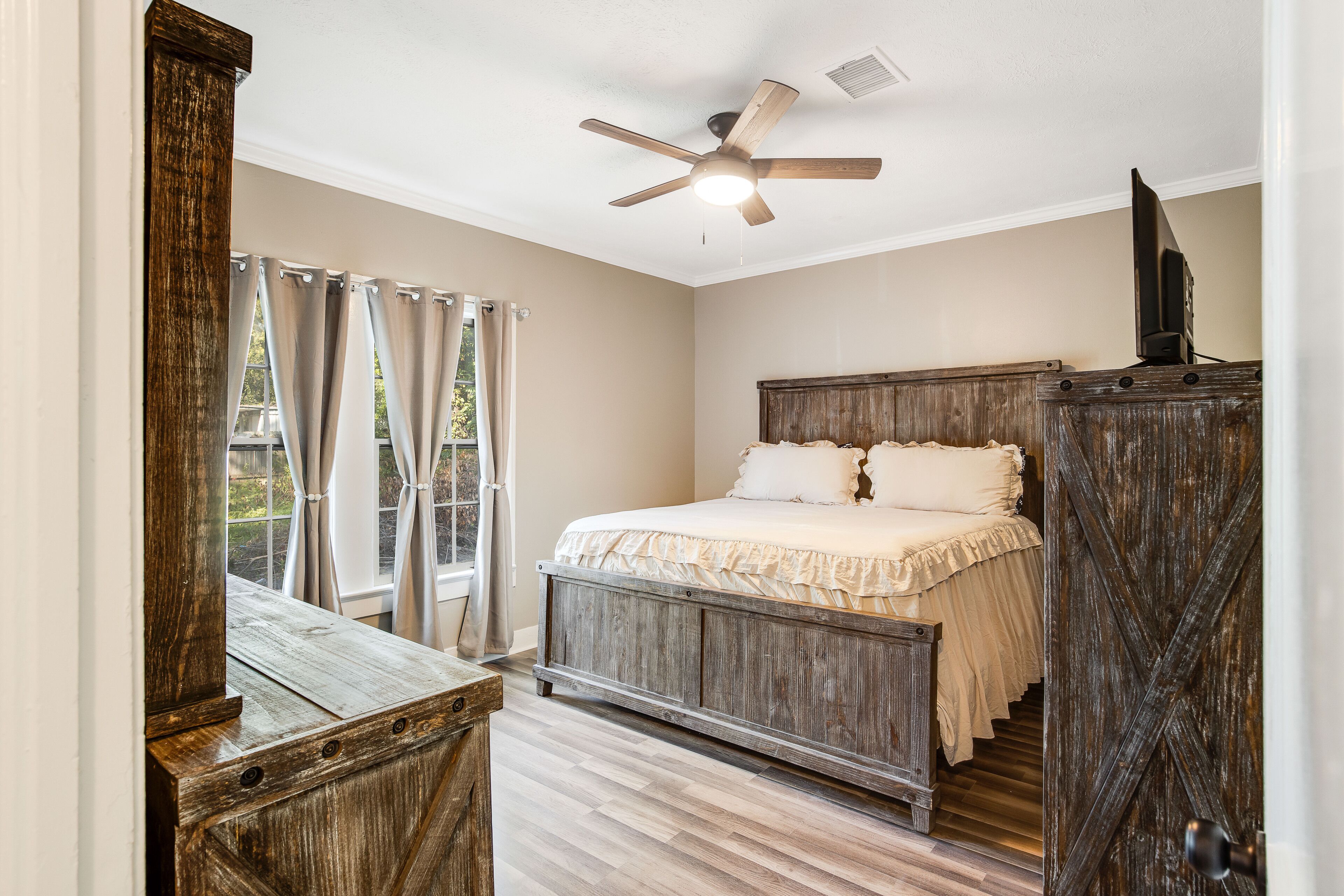 A bedroom with a wooden bed, a ceiling fan, and a view of the outdoors through curtains.