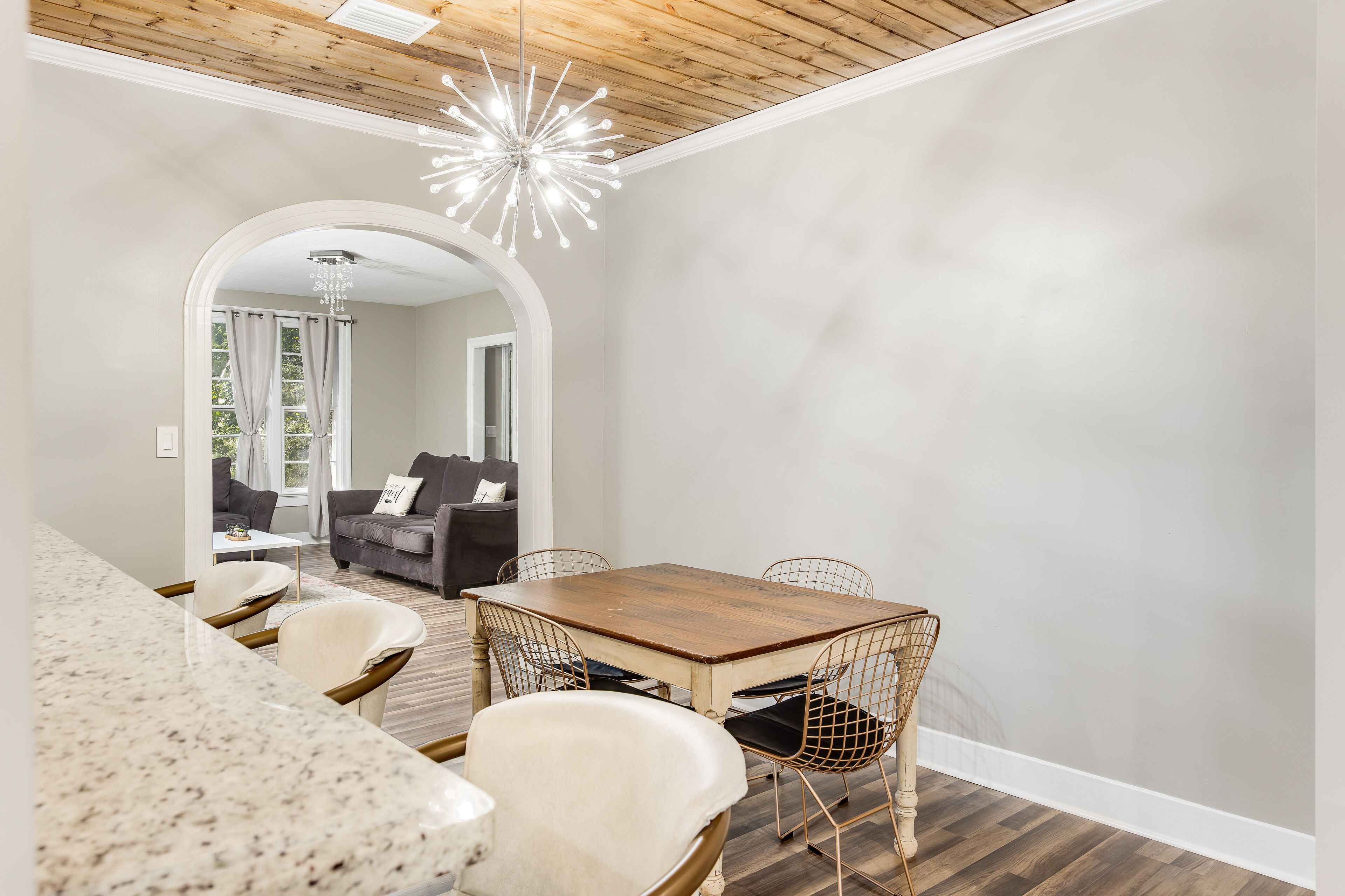 Un comedor con mesa de madera y sillas de alambre, una lámpara de araña y vista a una sala de estar con sofá y cortinas.