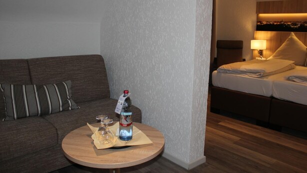 Habitación de hotel con sofá, una mesa redonda de madera, una botella y vasos, y una cama con mesita de noche.