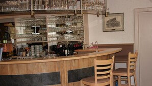 Una zona de bar con una barra curva, dos taburetes de madera y estantes llenos de copas y botellas.